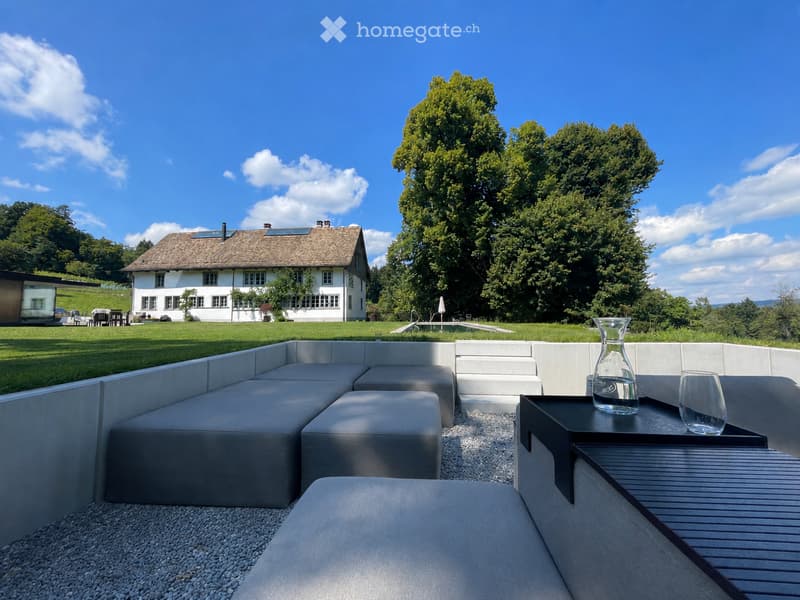 Modern outdoor seating area with grey sectional sofa, small table with glass decanter and glass, surrounded by gravel and white concrete walls, with a large house and swimming pool in the background.