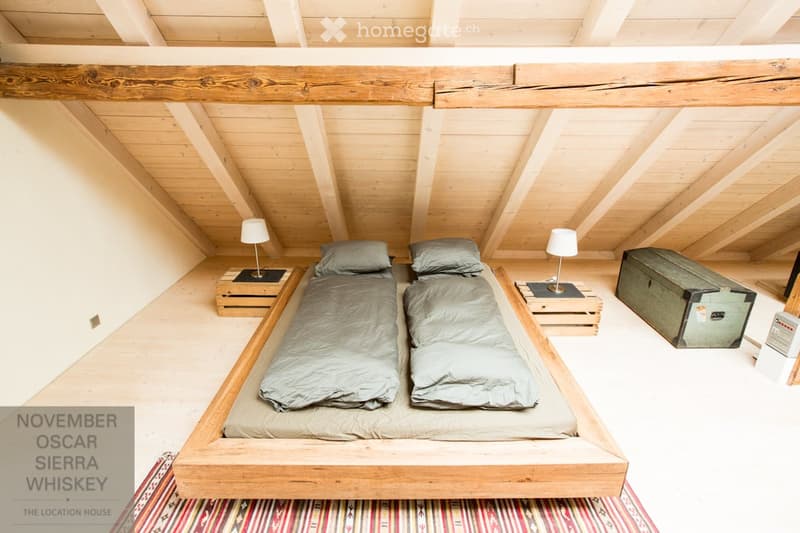 Cozy attic bedroom with wooden beams, a large bed with two sleeping bags, nightstands with lamps, and a vintage trunk.