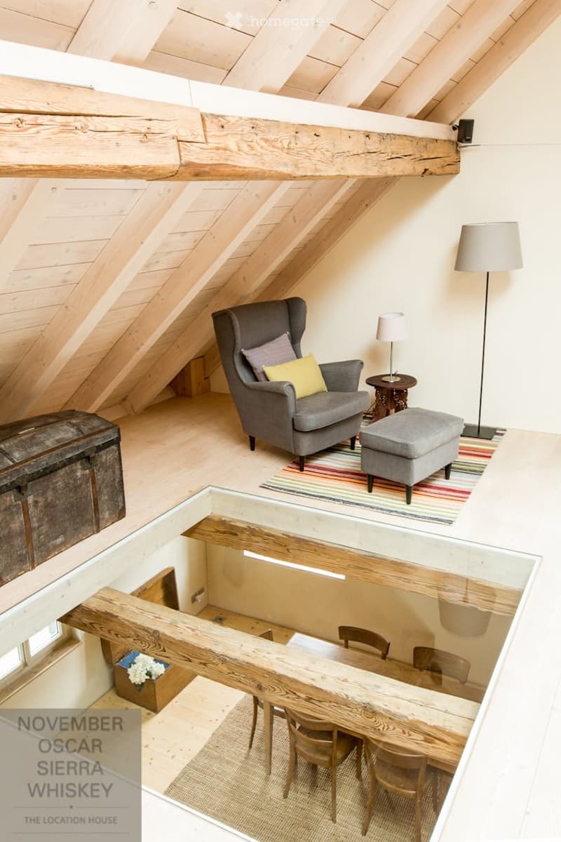 Loft-style living space with exposed wooden beams, a grey sofa with pillows, an ottoman, a small table lamp, a floor lamp, a wooden chest, and a visible dining area below with a wooden table and chairs.
