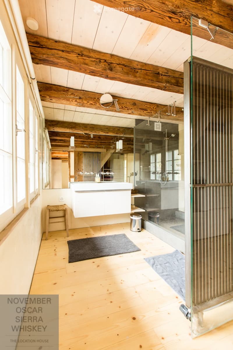 Bathroom with wooden ceiling beams, white sink, glass shower enclosure, and a small wooden stool near the sink.