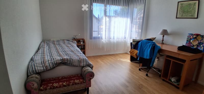 Cozy bedroom with wooden flooring, a bed with striped bedding, a small chair, a desk with a lamp, and a nightstand.