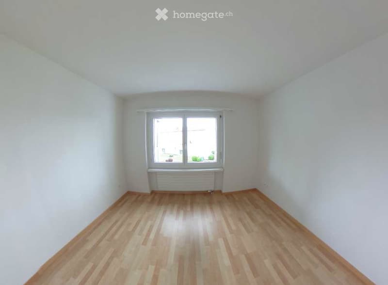 Empty room with light wooden flooring, white walls, and a single window with a radiator below it.