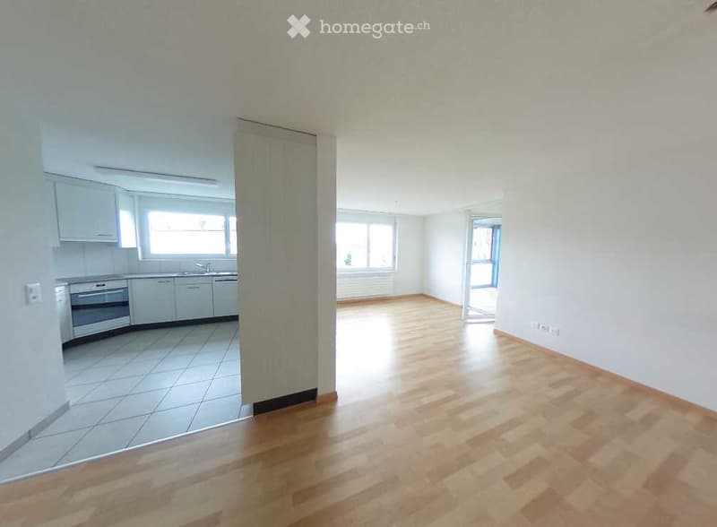 Empty room with wooden flooring and white walls, connected to a small kitchen area with white cabinets and appliances.