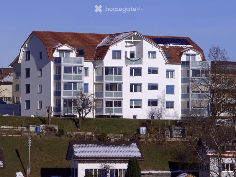 A multi-story residential building with white walls, large windows, and a red-tiled roof, featuring solar panels on the roof and a festive wreath on one of the balconies.