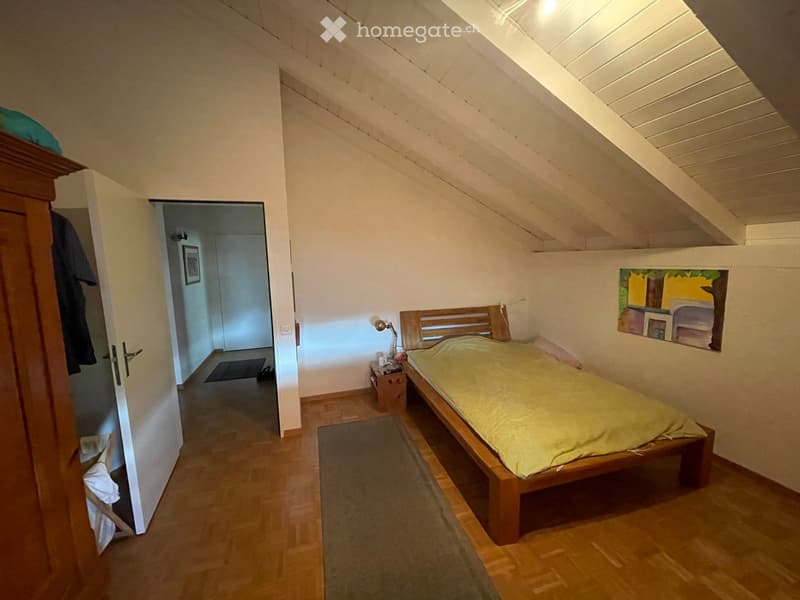 Cozy attic bedroom with wooden bed, nightstand, lamp, and painting on the wall. Slanted wooden ceiling and wooden flooring.