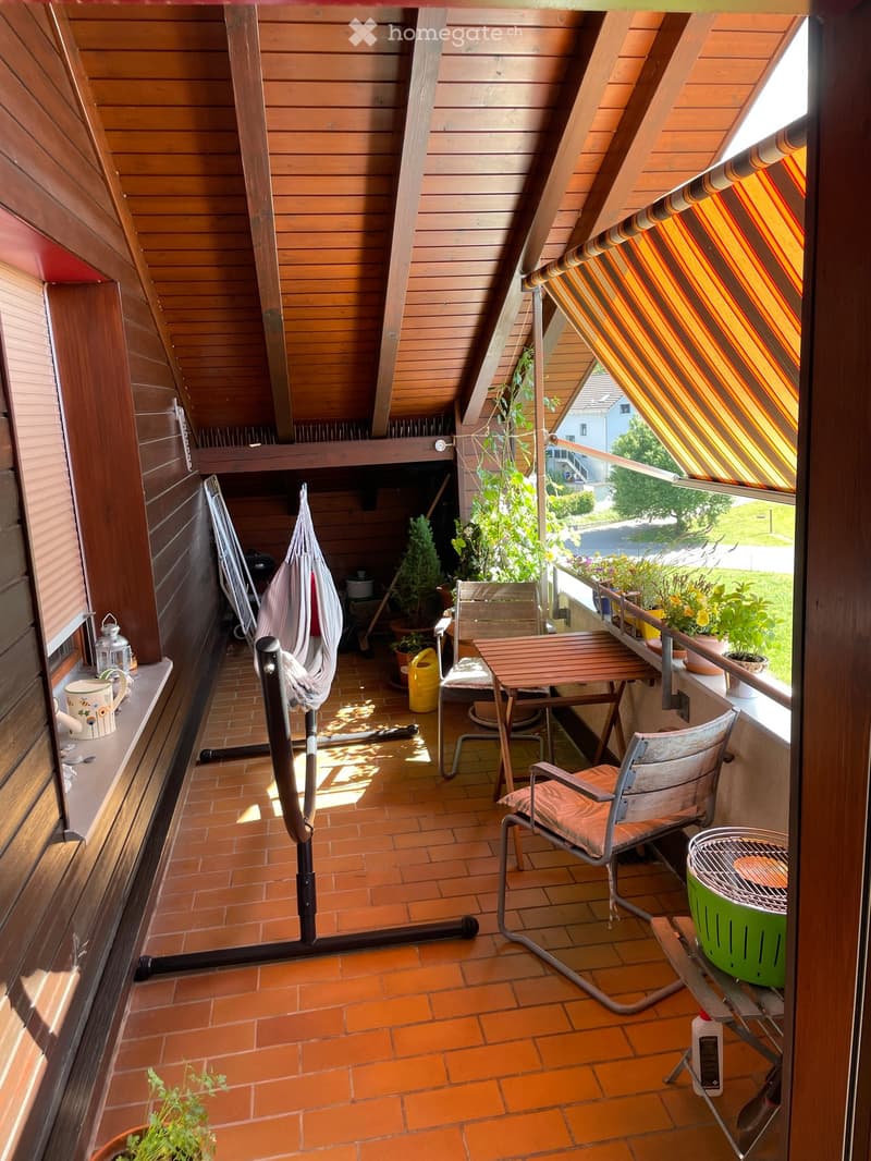 Wooden balcony with slanted roof, tiled floor, table, chairs, hammock, grill, and potted plants.