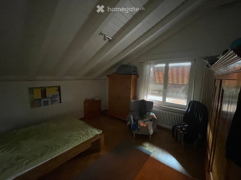 Cozy attic bedroom with wooden floors, slanted ceilings, and large windows. Features a bed, a chair with a blanket, a wardrobe, and a radiator.