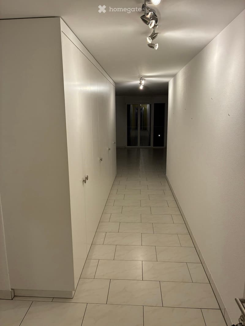 Empty hallway with tiled floor, white walls, and modern lighting fixtures. Features a closed door on the left and an open doorway at the end leading to another space.