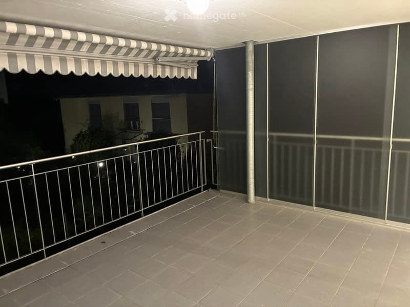 Balcony with tiled floor, metal railing, and retractable awning, overlooking a residential area at night.