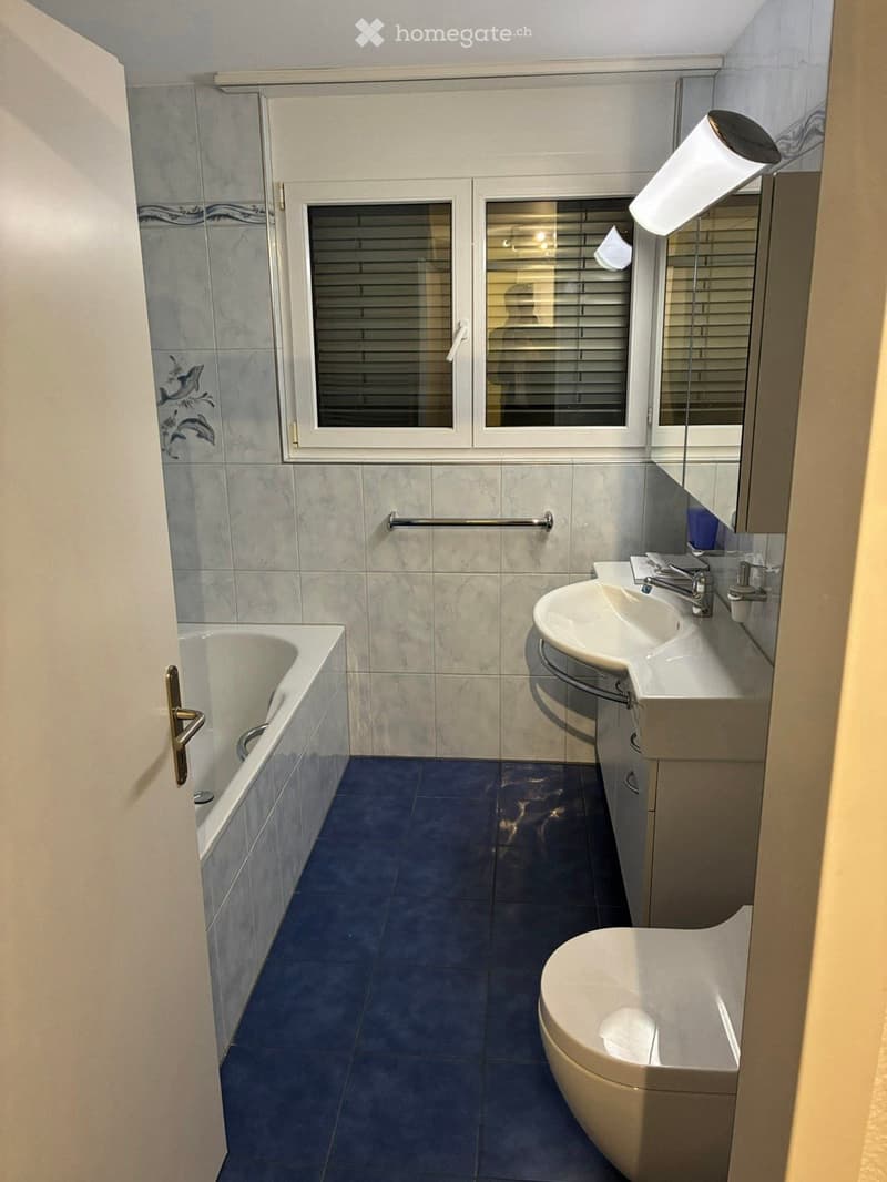 Bathroom with a bathtub, sink, toilet, mirror, and a window with blinds. The walls are tiled with a decorative pattern, and the floor is covered with dark blue tiles.