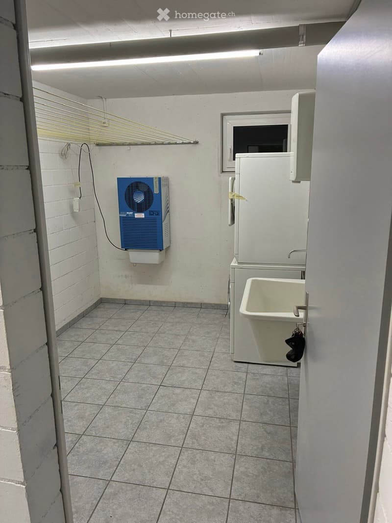 A small, tiled bathroom with a washing machine, a sink, and an air conditioner mounted on the wall. The space is clean and functional with a window providing natural light.