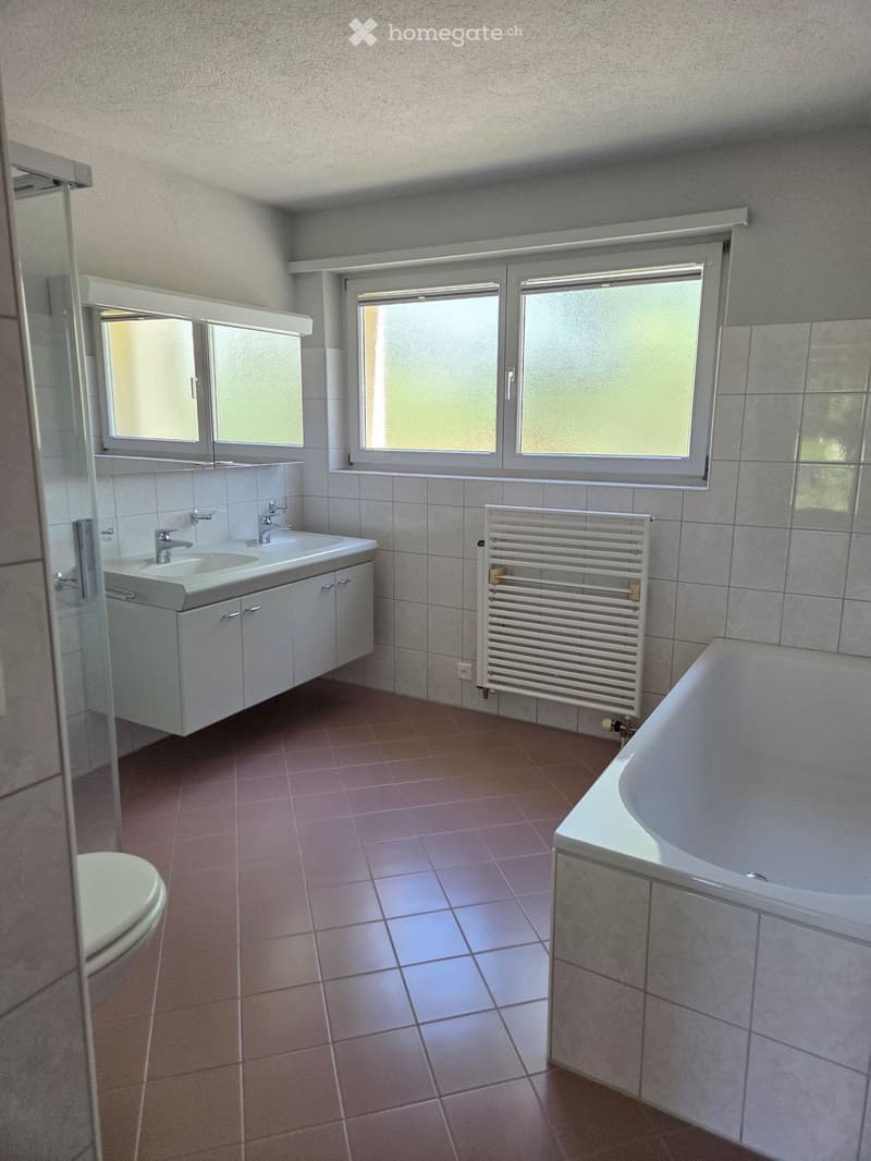 Modern bathroom with a bathtub, sink, toilet, radiator, and large windows. Features tiled walls and floor, and a mirrored cabinet above the sink.