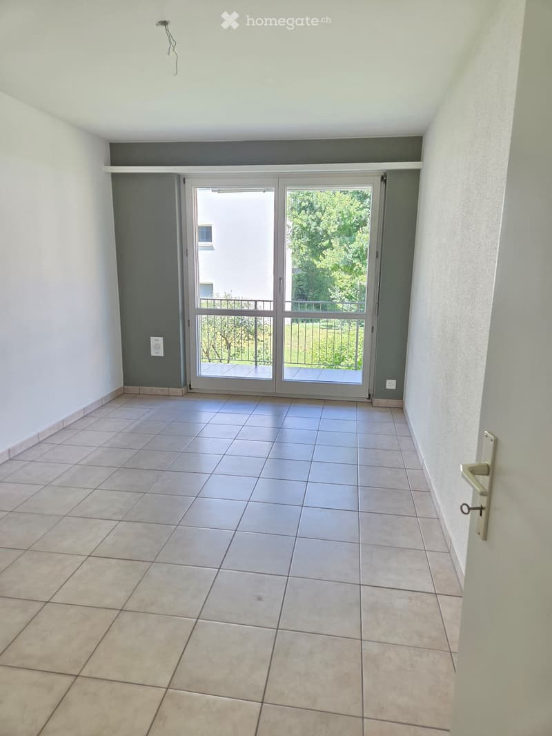 Empty room with tiled floor, white walls, and a sliding glass door leading to a balcony with a metal railing and greenery visible outside.