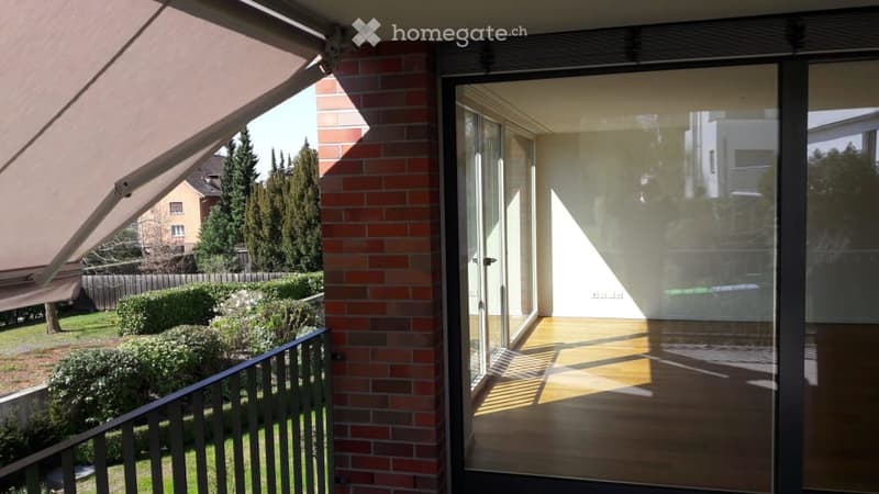 Balcony with wooden flooring, glass sliding doors, and a view of a garden with bushes and trees.