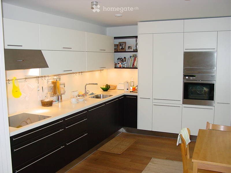 Modern kitchen with white cabinets, stainless steel appliances, and wooden flooring. Features include a sink, oven, and a small dining area with a table and chairs.