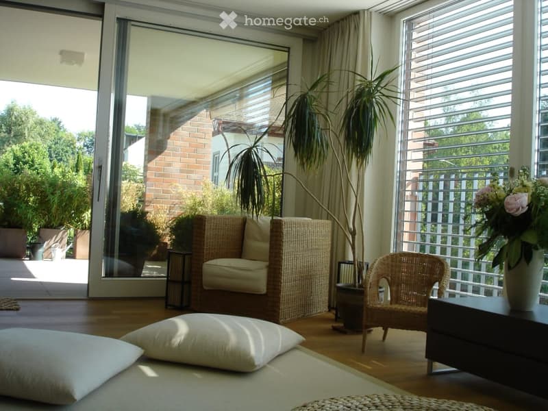 Bright living room with large windows and sliding glass doors leading to a patio. Features a wicker sofa, wicker chair, small table, potted plants, and a flower vase on a side table.
