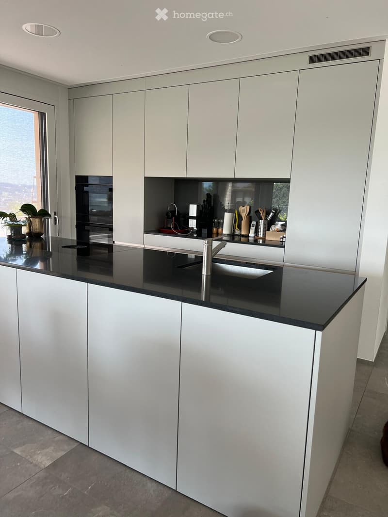 Modern kitchen with white cabinets, black countertop, stainless steel sink, and various appliances including an oven and fridge. The space is well-lit with recessed ceiling lights and features a large window with a view.