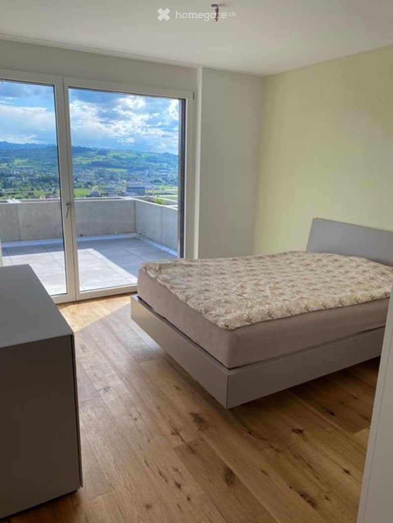 Modern bedroom with wooden flooring, large window offering a scenic mountain view, a grey bed with a textured duvet, and a matching nightstand.