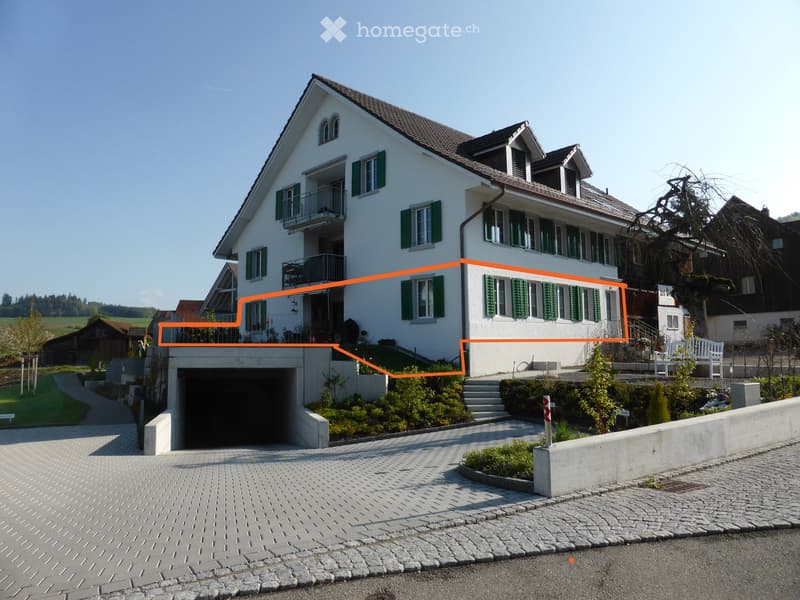 Two-story white house with green shutters, multiple windows, and a garage entrance. The house features a well-maintained garden and a paved driveway.