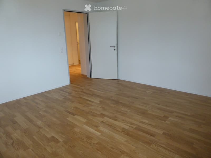 Empty room with wooden flooring and white walls, featuring a closed white door and an open doorway leading to another room.