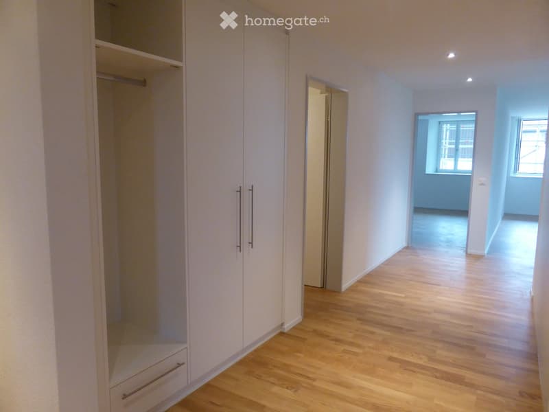 Empty hallway with wooden flooring, white walls, built-in closet with mirrored doors, and two open doorways leading to other rooms.