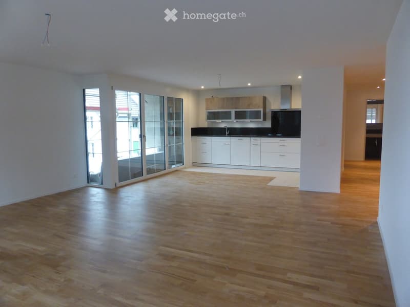 Empty room with wooden flooring, white walls, and a large glass door leading to a balcony. The kitchen area is visible in the background with white cabinets and stainless steel appliances.
