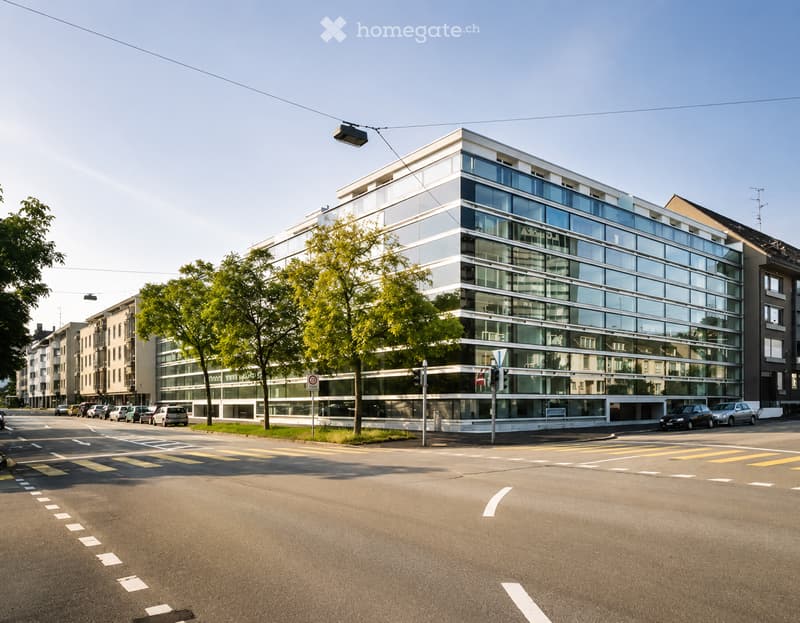Modern multi-story building with large glass windows and a clean, contemporary design, situated at a street corner with trees and parked cars nearby.