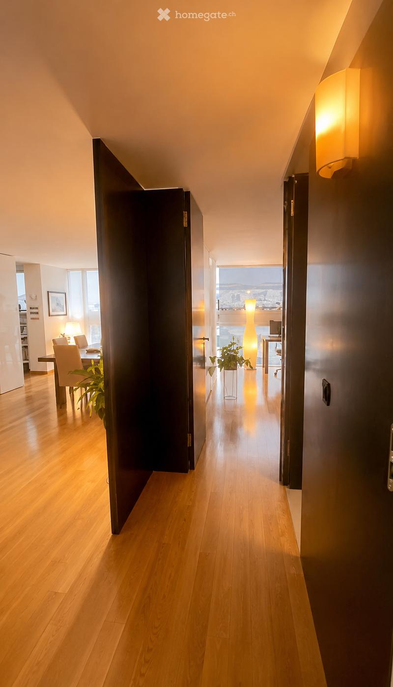 Modern hallway with wooden flooring, warm lighting, and a view of the cityscape through a large window at the end.