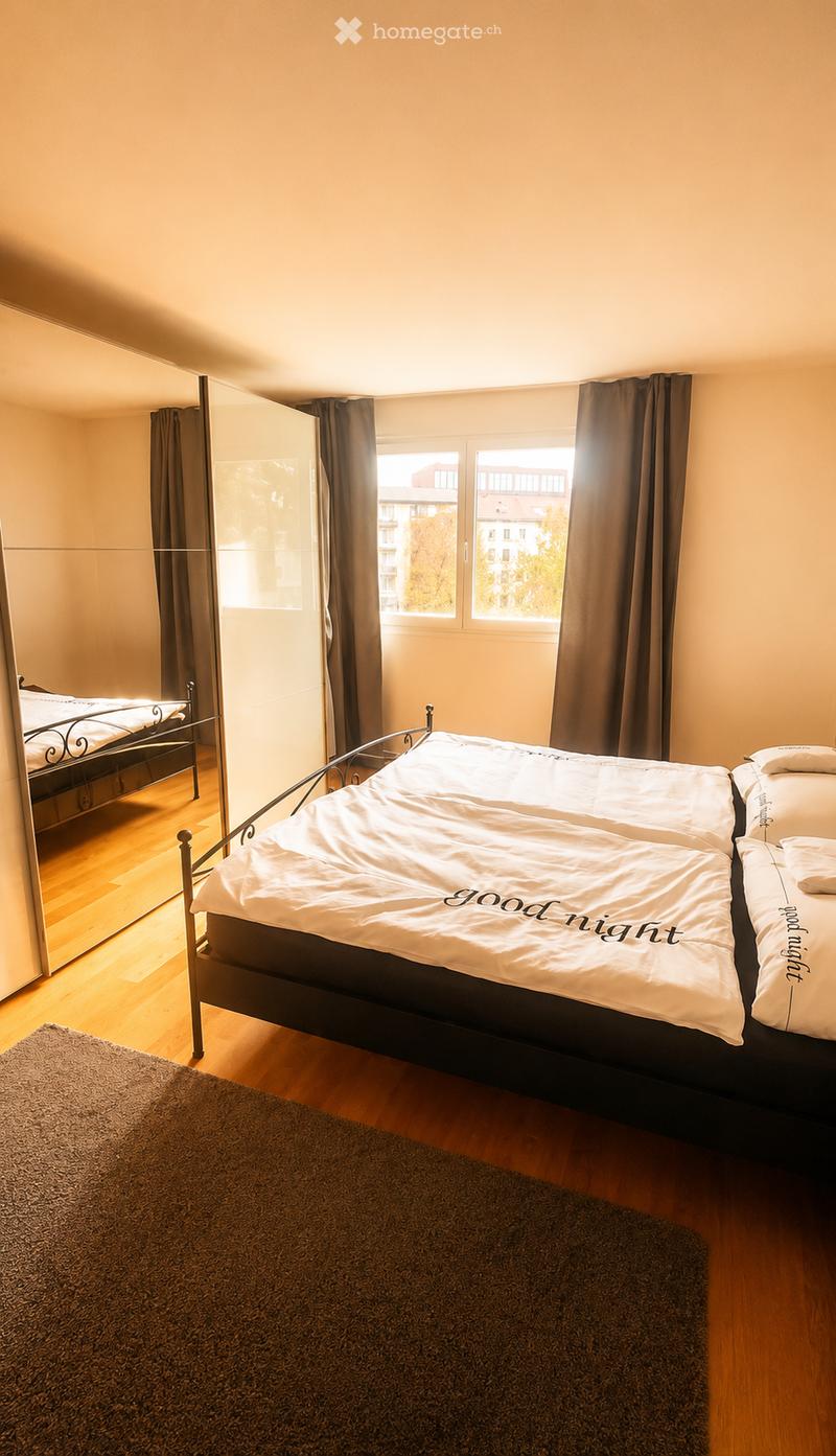 Cozy bedroom with wooden flooring, large window with curtains, double bed with white bedding, and a mirror on the wall.