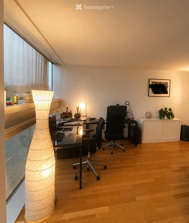 Modern office space with wooden flooring, large window, desk with laptop, chairs, and decorative lamp.