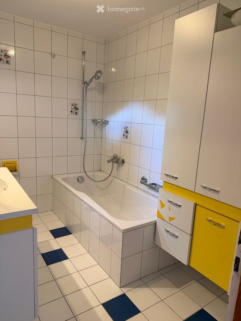 Modern bathroom with white tiles, a bathtub, a showerhead, and white cabinets with yellow accents.