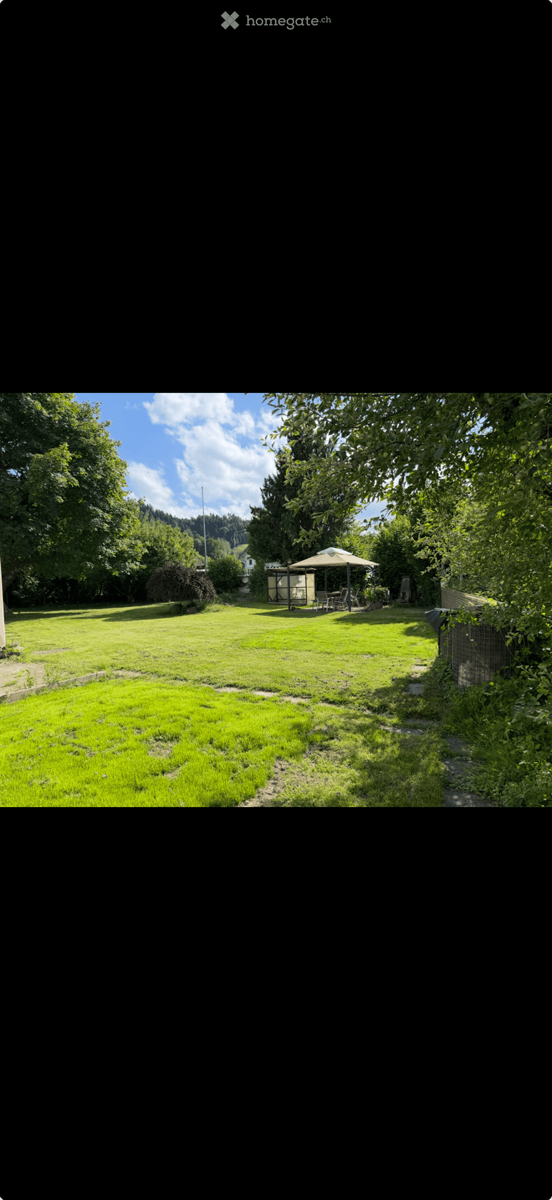 A spacious, well-maintained garden with a green lawn, trees, and a small outdoor seating area with a table and chairs under a canopy.