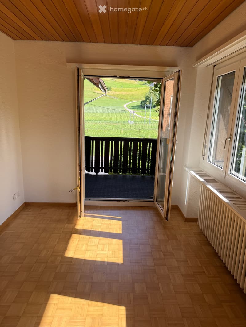 Empty room with wooden flooring, white walls, and a large open sliding door leading to a balcony with a view of a green lawn and trees.