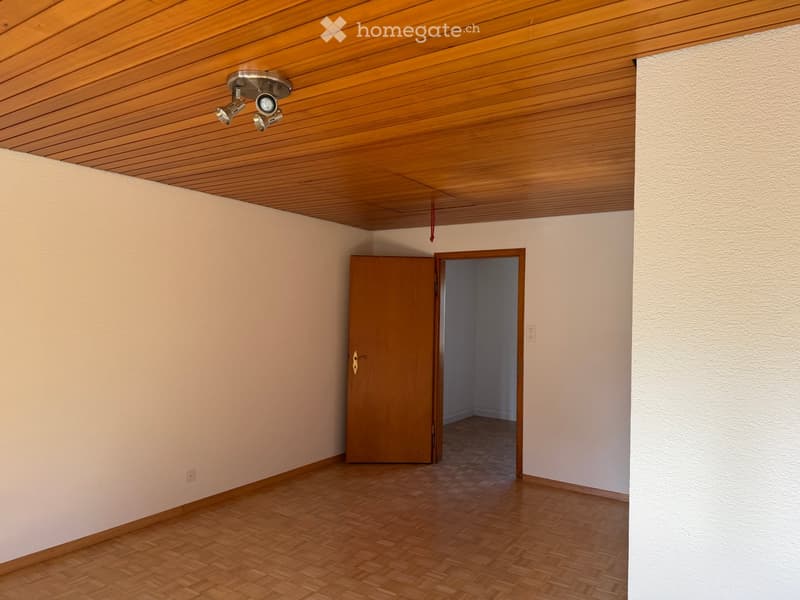 Empty room with wooden flooring, white walls, and a wooden door leading to another space. The ceiling features wooden paneling and recessed lighting.