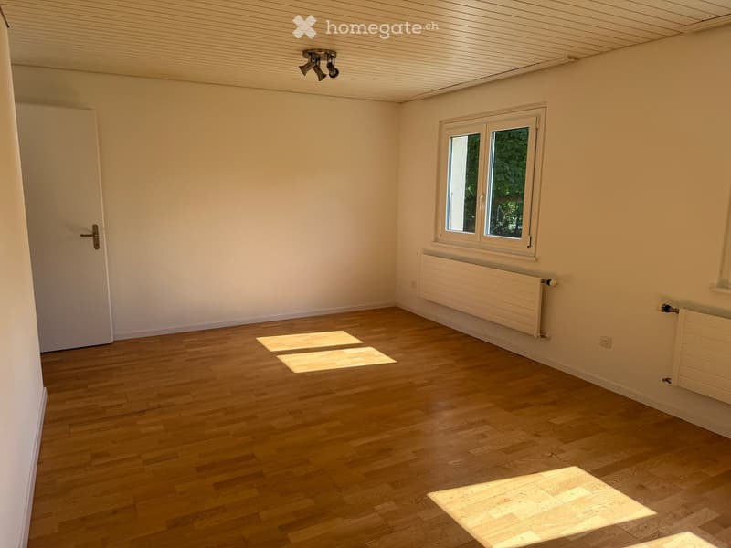 Empty room with wooden flooring, white walls, a single window, and a door. Natural light streams in, casting shadows on the floor.