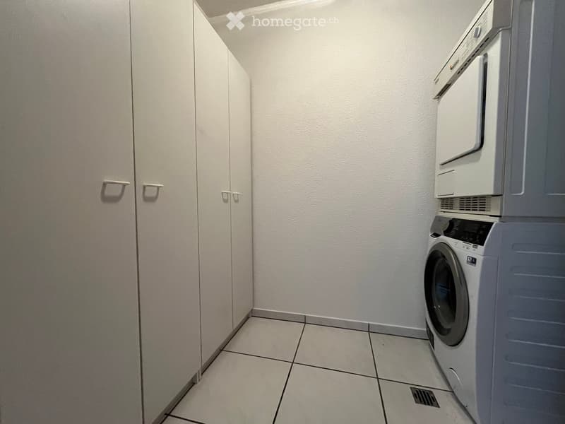 A small, tiled room with a washing machine and dryer stacked vertically, accompanied by a white cabinet with silver handles.