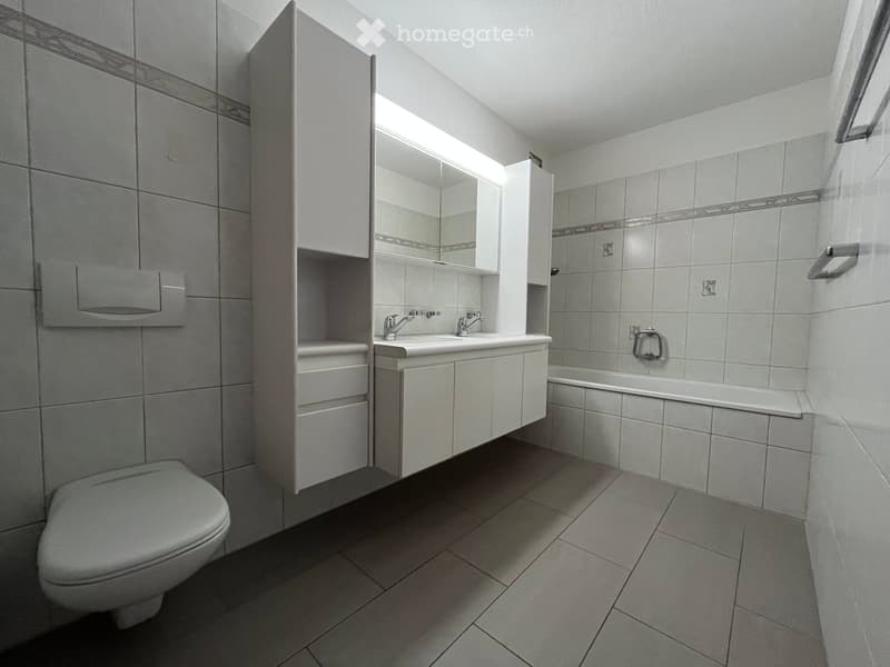Modern bathroom with white tiles, a toilet, a double sink with cabinets, a bathtub, and a mirror above the sink.