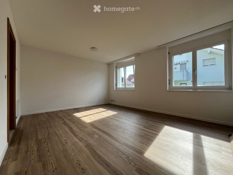 Empty room with wooden flooring, white walls, and large windows allowing natural light to enter.