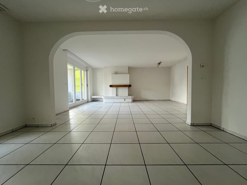 Empty room with white tiled floor, white walls, and a fireplace with a wooden mantelpiece. Large windows and an arched doorway are visible.