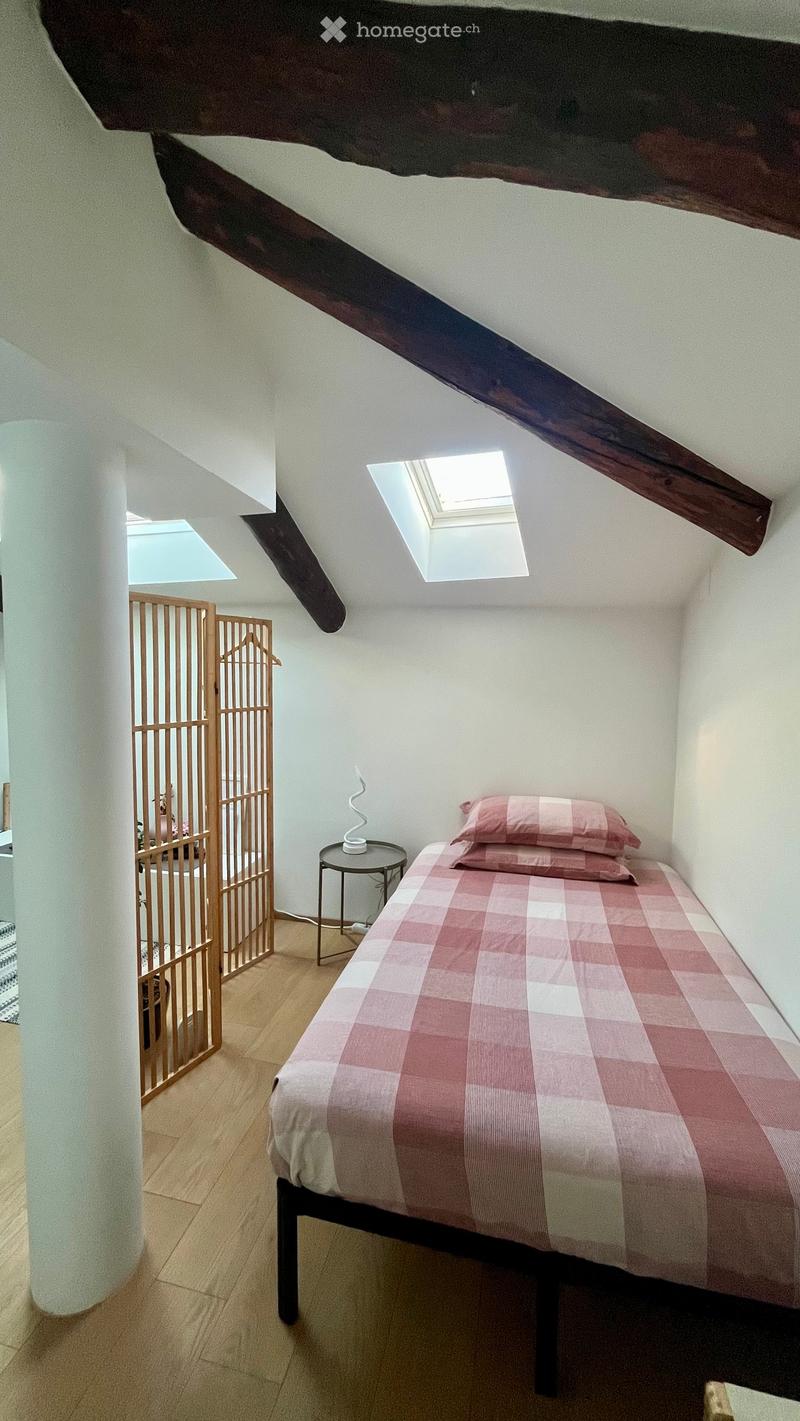 Cozy attic bedroom with wooden slanted ceiling beams, skylight window, bed with pink checkered bedding, small round nightstand with lamp, and a wooden screen partition.