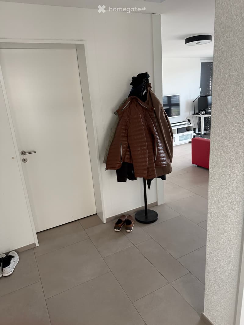 A hallway with a coat rack holding jackets, a pair of shoes, and a small red stool. A white door is visible on the left, and a TV screen is partially visible in the background.