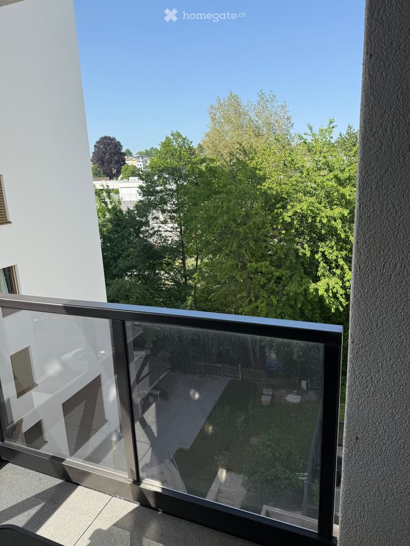 Balcony with glass railing, overlooking a green area with trees and a neighboring building.