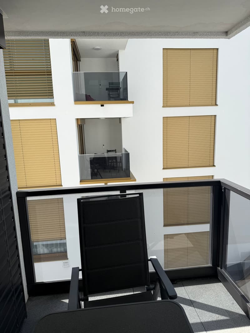Balcony with modern glass railings, several windows with blinds, and a single black chair.