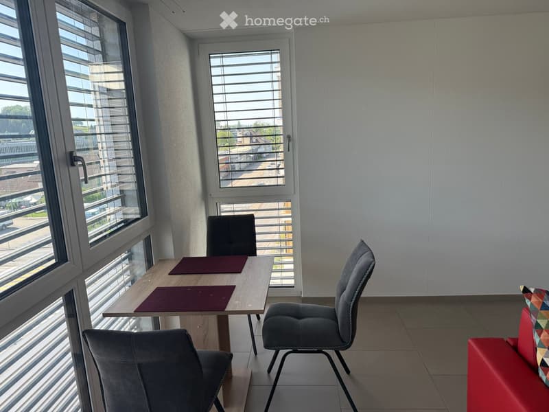 A modern, furnished room with large windows, a red sofa, a wooden table, and two grey chairs.