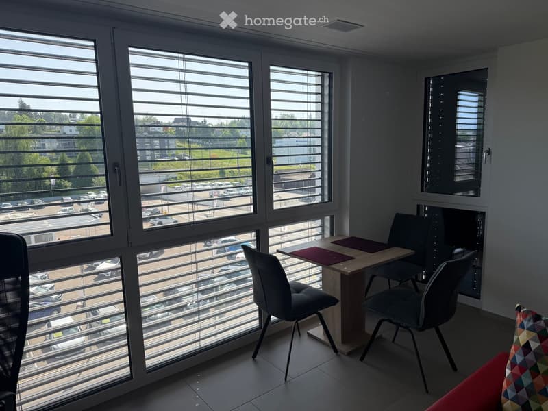 Office space with large windows, a small wooden table, two chairs, and a colorful cushion on the floor.