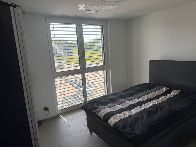 Modern bedroom with a large bed, dark bedding, and large windows with blinds overlooking a parking area.