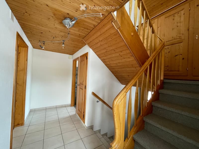 Wooden staircase with a handrail and carpeted steps, leading to a wooden door on the left and a wooden cabinet on the right. The ceiling is wooden with a modern light fixture.