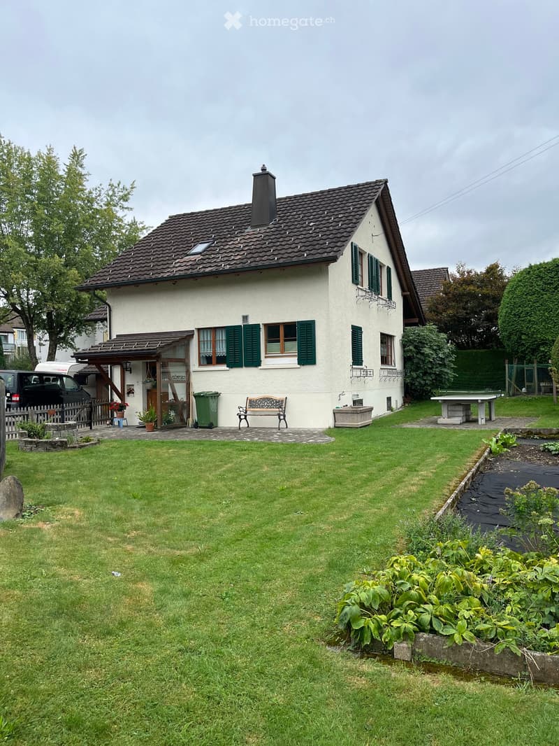 A two-story house with a well-maintained garden, featuring a lawn, a bench, a small table, and various plants and trees.