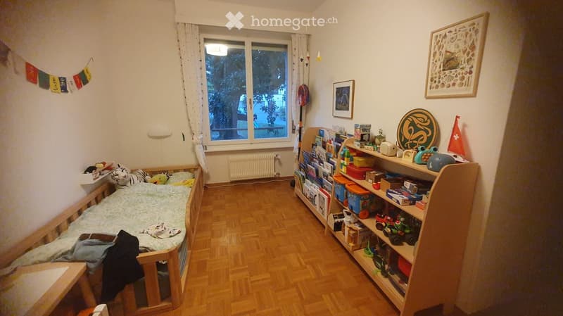 Child's bedroom with a small bed, bookshelf filled with books and toys, and a window with curtains.