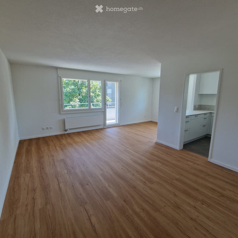 Empty room with wooden flooring, white walls, large windows, and a radiator. The room is unfurnished and has a door leading to another area.